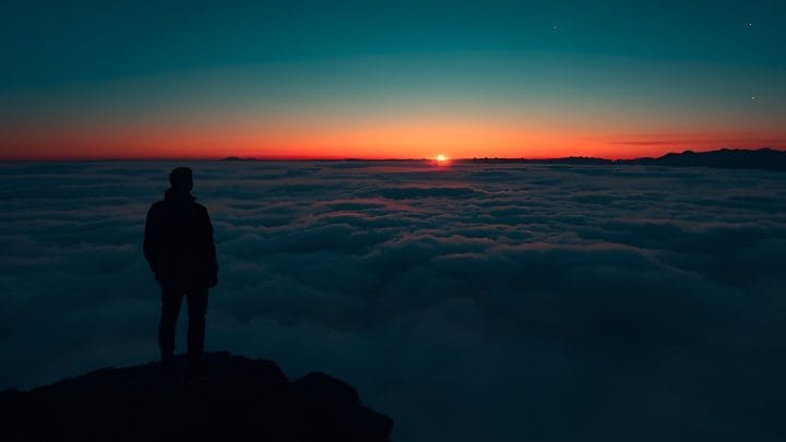 A lone figure stands silhouetted on a rocky summit above a vast sea of fog, facing a deep red and teal sunrise on the horizon — representing clarity, ambition, and rising above the noise of brain fog.