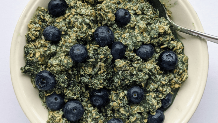Overhead view of a bowl of green overnight oats with greens powder, chia seeds and fresh blueberries on top, spoon resting in the bowl